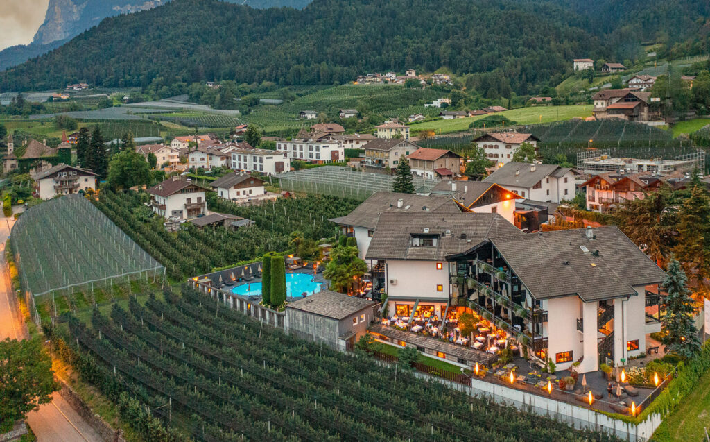 Yoga Retreat Südtirol: Eine luftige Außenansicht eines luxuriösen Hotels in Südtirol bei Abenddämmerung, geschmückt mit warmen Lichtern, die eine einladende Atmosphäre schaffen. Der Außenpool und die umliegenden Sitzbereiche sind belebt mit Gästen, umgeben von weitläufigen Weinbergen und der sanften Silhouette der Südtiroler Alpen im Hintergrund, ideal für einen erholsamen Yoga-Retreat.