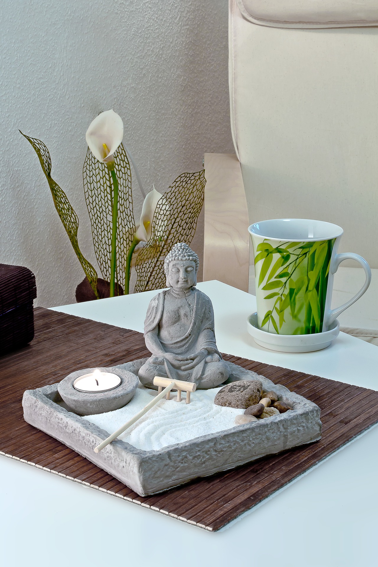 Ein Zen-Dekorationsset auf einem Tisch mit einer Buddha-Statue im Zentrum, umgeben von einer brennenden Kerze, Räucherstäbchen und sanft geschwungenen Calla-Lilien im Hintergrund, daneben steht eine Tasse mit Bambusmotiv. Dieses stimmungsvolle Arrangement verkörpert die ruhige Atmosphäre, die für Yin Yoga online Kurse typisch ist.