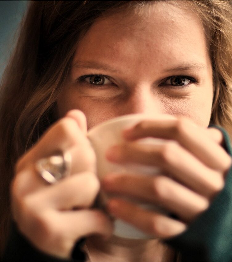Eine junge Frau blickt über den Rand einer Tasse, die sie mit beiden Händen hält, direkt in die Kamera, während ein warmes Lächeln ihr Gesicht umspielt. Dieses Bild könnte in einem Kontext für einen Artikel über die beruhigende Wirkung von Yin Yoga online verwendet werden, wo eine Tasse Tee die Atmosphäre der Entspannung unterstreicht.
