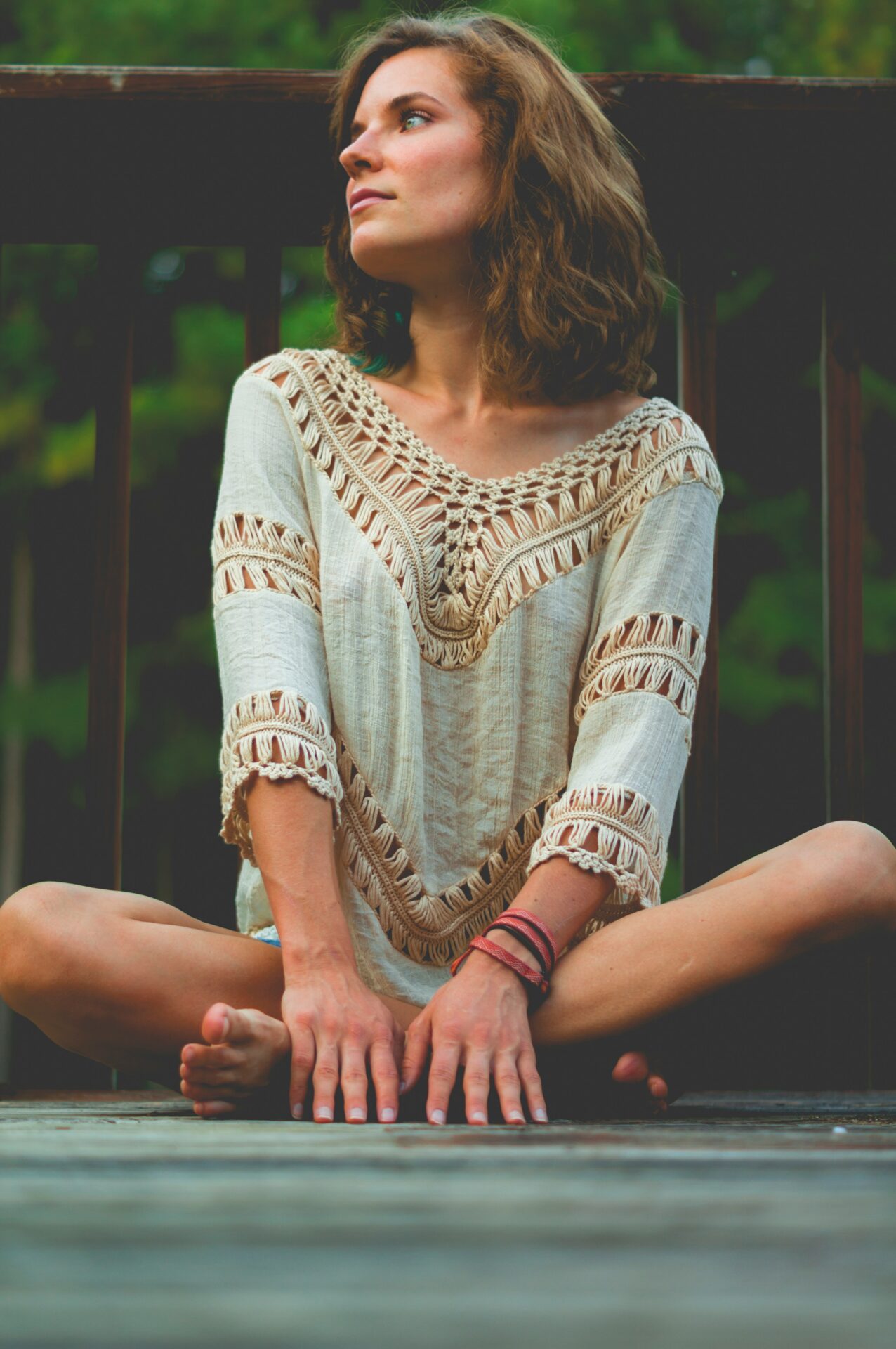 Eine Frau in einer entspannten Yin-Yoga-Pose sitzt im Lotussitz auf einer Holzterrasse, gekleidet in ein beiges, makrameeartiges Top, während sie gelassen in die Ferne schaut, eine Einladung zur Ruhe und Entspannung in einem Yin Yoga Online-Kurs.