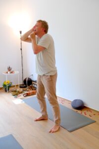 Yin Yoga Ausbildung Hamburg: Kursleiter Jan Wolk demonstriert Gesichtsübung in hellem Yogastudio mit Yogamatte, Meditationskissen und Entspannungszubehör