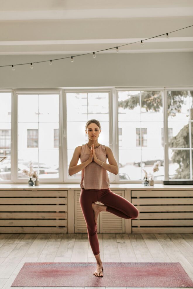 Frau praktiziert Yoga im Baum-Asana (Vrikshasana) in hellem Studio mit großen Fenstern im Hamburger Osten, trägt rosa Top und rote Leggings