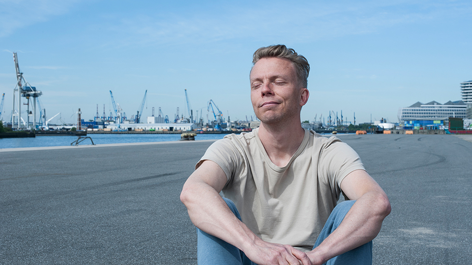 Jan Wolk sitzt mit geschlossenen Augen vor dem Hamburger Hafen
